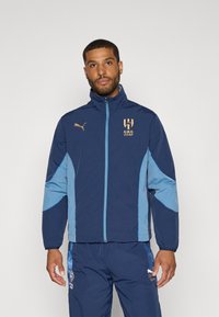 Chaqueta azul marino con acentos en azul claro, hecha de material liviano. Presenta un cuello alto, cierre de cremallera y logo bordado en el pecho.