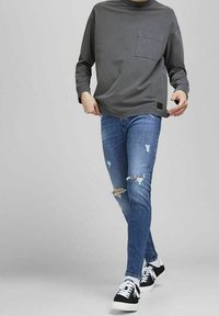 Maglietta grigia a maniche lunghe con taschino sul petto, abbinata a jeans blu aderenti con dettagli usurati e sneaker nere con suola bianca.