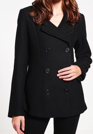 Nainen, jolla on musta kaksirivinen takki, jossa on kuusi nappia, seisoo käsi vyötäröllä ja toinen rentona sivullaan.