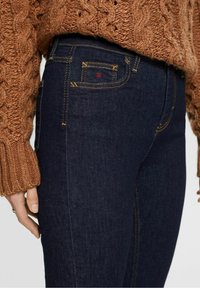 Jeans de mezclilla azul oscuro con un diseño ajustado. Presentan costuras amarillas y un pequeño detalle de logo rojo en el bolsillo trasero. Suéter marrón texturizado encima.