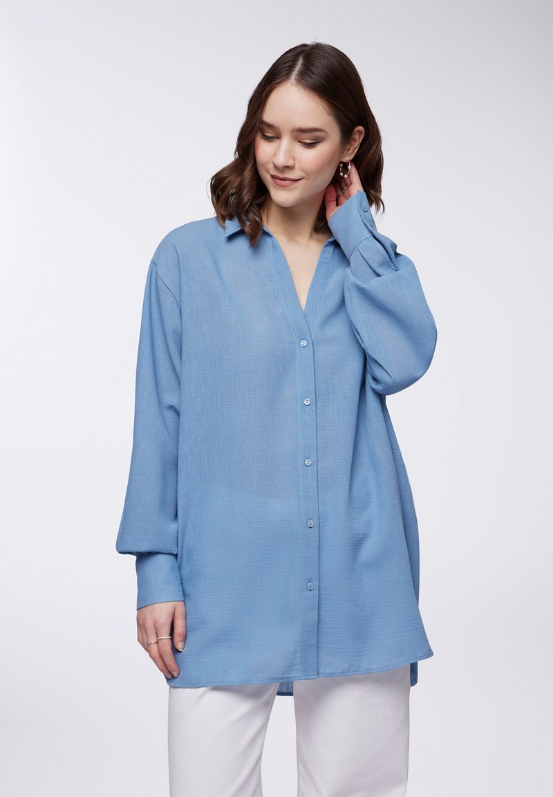Camicia button-up blu chiaro realizzata in tessuto morbido, con scollo a V, maniche lunghe e vestibilità rilassata. Texture liscia con un leggero lucido.