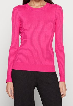 Femme portant un pull à manches longues côtelé rose vif assorti d'un pantalon noir texturé, debout devant un fond uni.