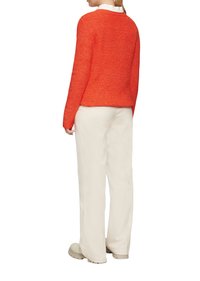 Oranger Strickpullover mit strukturierter Oberfläche, Rundhalsausschnitt und langen Ärmeln, kombiniert mit weißen, weiten Hosen und beigen Schuhen.