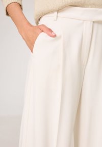 Pantalons blancs ajustés avec une texture lisse, présentant des poches latérales et des plis nets, assortis à un pull en tricot beige clair.