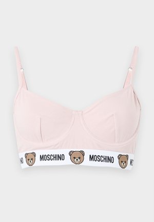 Vrouw staat blootsvoets, draagt een roze beha met berenlogo en wit Moschino-ondergoed, kijkt naar links tegen een effen achtergrond.