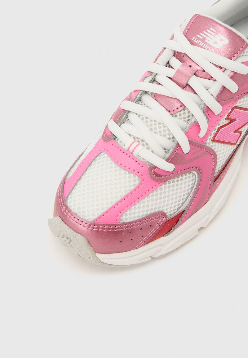 Chaussure de course New Balance blanche et rose avec des lacets blancs, une tige en mesh et des surcouches métalliques roses sur un fond blanc.