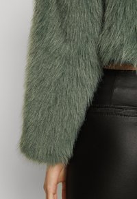 Grøn faux fur jakke med lange ærmer, der har en blød tekstur og cropped design, hvisettet med sorte tætsiddende bukser.