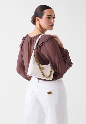 Vrouw met donker haar in een knot draagt gouden oorringen, een mauve blouse met ruches, witte broek en heeft een witte tas met een gouden kettingriem bij zich.