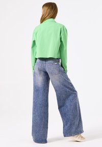 Grünes, kurzes Hemd aus leichtem Stoff, mit langen Ärmeln. Weit geschnittene, ausgewaschene blaue Jeans aus Denim, mit Gesäßtaschen und sichtbaren Schäden, neutrale Schuhe.