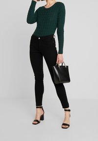 Suéter verde de punto acanalado, jeans ajustados negros, sandalias negras con tacón, sosteniendo un bolso negro pequeño y estructurado con detalles dorados.