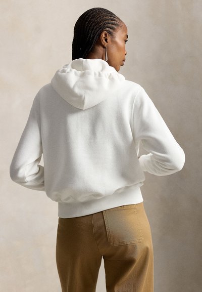 Woman with braided hair wearing a white hooded sweatshirt and brown pants, standing with hands on hips against a plain background.