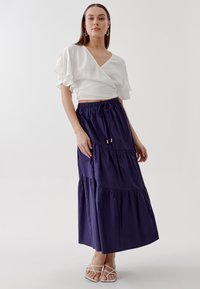 Top bianco avvolgente con maniche a volant abbinato a una gonna maxi blu navy a strati. Presenta una vita con coulisse e dettagli in oro.