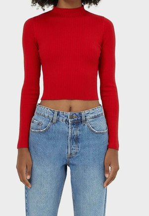 Femme portant un crop top ajusté à manches longues rouge côtelé, associé à un jean en denim bleu clair taille haute, sur un fond uni.