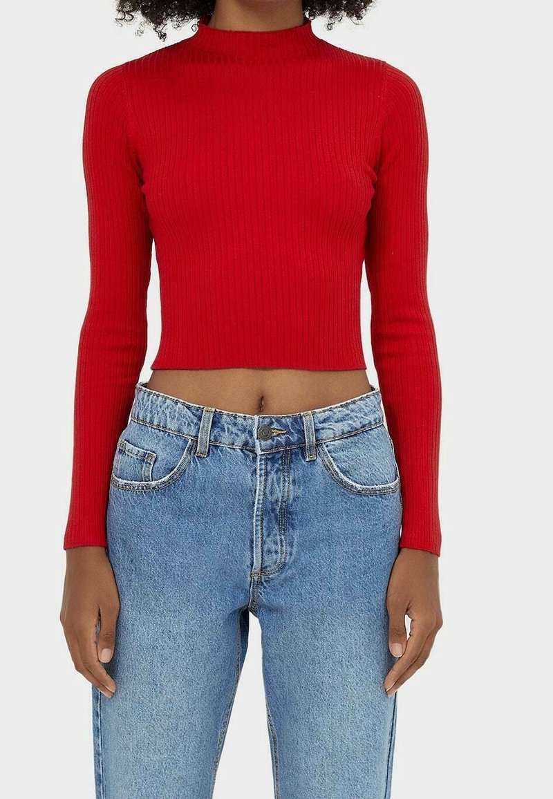 Femme portant un crop top ajusté à manches longues rouge côtelé, associé à un jean en denim bleu clair taille haute, sur un fond uni.