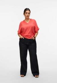 Blusa coral de manga corta y cuello en V combinada con pantalones vaqueros oscuros de campana. La blusa tiene un corte holgado; los pantalones presentan una textura de denim clásica.