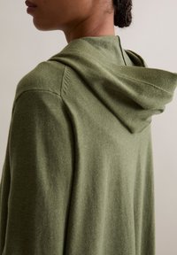 Vêtement à capuche couleur vert olive, fabriqué en tissu doux, avec une coupe décontractée, des détails de couture visibles et un design simple et minimaliste.