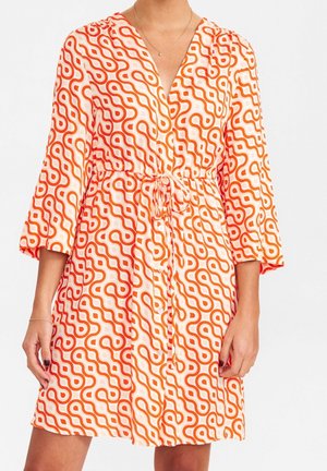 Femme portant une robe mi-longue avec des motifs de lignes ondulées orange, manches trois-quarts, ceinture nouée devant et fermeture à boutons.