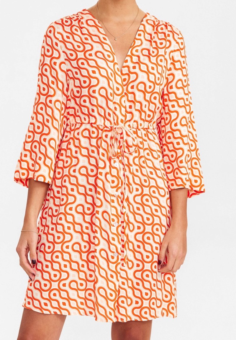Femme portant une robe mi-longue avec des motifs de lignes ondulées orange, manches trois-quarts, ceinture nouée devant et fermeture à boutons.