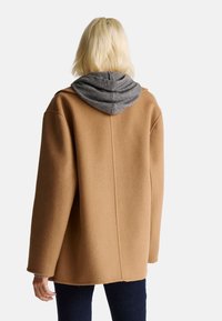 Hnědý kabát vyrobený z vlněné směsi, pod kterým se nachází šedá kapuce, kleslá ramena a uvolněný střih bez viditelných zapínání.