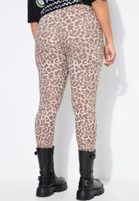 Osoba oblečená v béžových a hnědých legínách s leopardím potiskem zastrčených do černých masivních polokozaček, stojící na bílém pozadí.