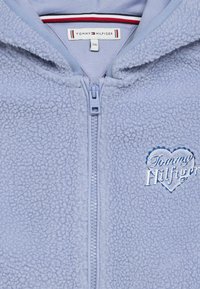 Svetlomodrá fleecová bunda na zips s vyšívaným logom v tvare srdca. Štruktúrovaná tkanina a kontrastná podšívka na golieri.