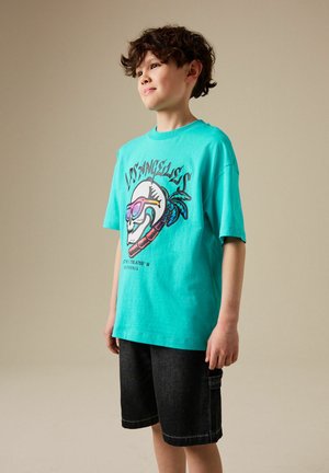 Băiat cu păr creț purtând un tricou turcoaz cu grafica Los Angeles și pantaloni scurți negri, stând pe un fundal bej simplu.