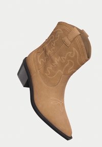 Bottine western en suède beige avec des coutures décoratives, des poignées de tirage, un embout pointu et un talon noir empilé.