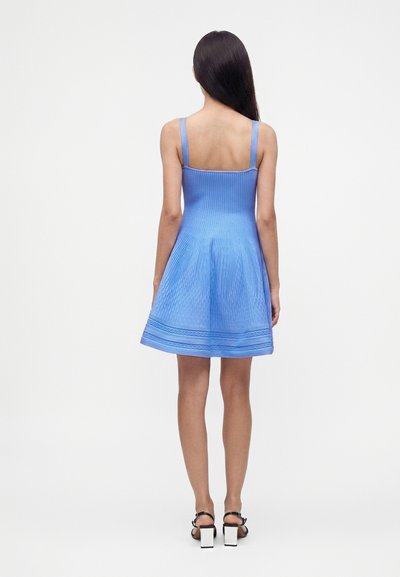Femme aux longs cheveux foncés porte une robe tricotée bleue sans manches et des sandales à brides noires avec des talons carrés blancs, debout de dos.