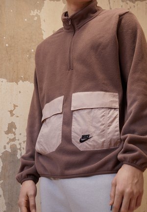 Osoba ubrana w brązowy polar z dwoma dużymi beżowymi kieszeniami z przodu oraz logo Nike na prawej kieszeni, stojąca na tle teksturowanej ściany.