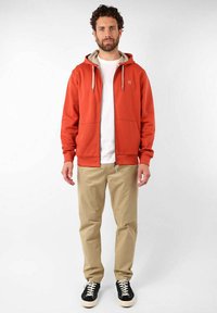 Haut à capuche zippé rouge avec doublure beige, capuche à cordon de serrage et poches avant, associé à un pantalon beige et des chaussures noires avec des lacets blancs.