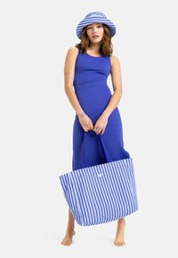 Grote gestreepte draagtas in blauw en wit, gemaakt van duurzaam materiaal. Het model draagt een effen blauwe jurk en een bijpassende gestreepte hoed.