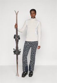 Ski-outfit met een crèmekleurig ritsfleece top en zwart-witte geblokte skibroek, met ski's in de hand en zwarte skischoenen aan.