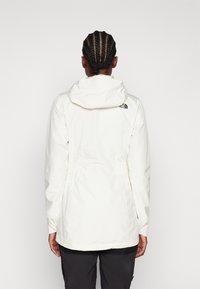 Giacca impermeabile bianca con cappuccio, dettaglio elastico in vita e logo sul retro; si distinguono la texture liscia e la vestibilità sagomata.