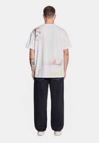 Biała graficzna koszulka z nadrukiem w kolorach czerwonym, żółtym i czarnym, zestawiona z luźnymi ciemnymi dżinsami i beżowymi sneakersami. Widok z tyłu.