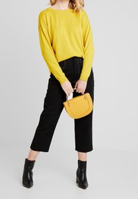 Geel gebreid sweater, zwarte cropped broek, zwarte puntige enkellaarsjes, houdt een kleine gele handtas vast met een gebogen bovenkant en zilveren hardware.