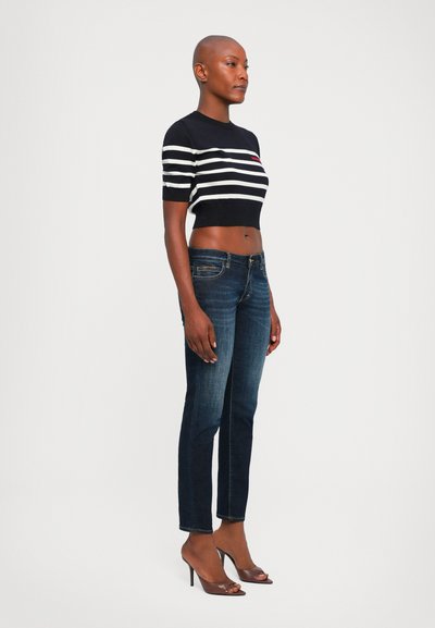 Woman standing sideways wearing a black and white striped cropped sweater, dark blue jeans, and brown high-heeled sandals on a white background.
