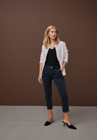 Šviesiaplaukė moteris stovi prie rudos sienos, dėvi baltą atverstą marškinių dalį, juodą V formos kaklo viršutinę dalį, tamsiai mėlynas trumpintas kelnes, juodus aukštakulnius mule ir sidabrinį apyrankę.