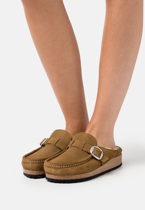 Brown suede clogs with a buckle, featuring a cushioned footbed and a cork outsole. Rounded toe and soft texture.