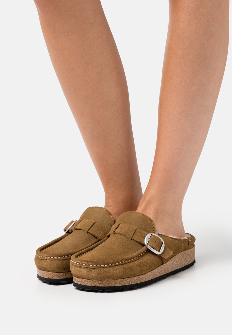 Zoccoli in suede marrone con fibbia, dotati di una soletta imbottita e una suola in sughero. Punta arrotondata e texture morbida.