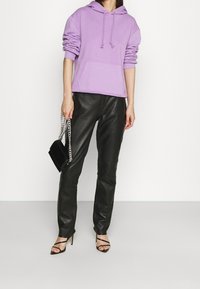 Lilafarbenes Kapuzensweatshirt mit einer Fronttasche, kombiniert mit schwarzen Lederhosen. Sie hält eine schwarze Handtasche mit einem silbernen Kettenriemen.