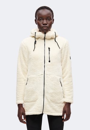Femme portant une veste en polaire couleur crème avec fermeture éclair devant, poche poitrine, capuche et poches pour les mains sur un fond clair uni.