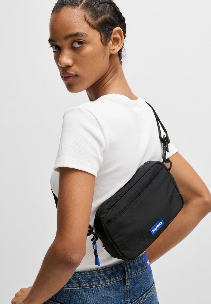 Bolso negro de hombro con textura acolchada, acento de logo azul, cierre de cremallera y correa ajustable, llevado sobre una camisa blanca y jeans de mezclilla.