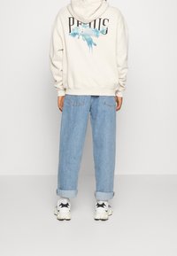 Krémová mikina s grafickým potiskem a modrými akvarelovými detaily na zádech, spárovaná s světle modrými volnými džíny a černo-bílými teniskami.