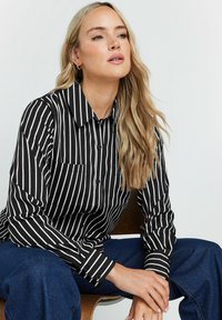 Camicia a righe bianche e nere con bottoni, maniche lunghe, design con colletto e una tasca sul petto. Tessuto liscio con una vestibilità su misura.