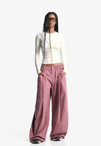 Top alb cu mâneci lungi, combinat cu pantaloni largi pal roz deschis, cu talie cu șnur și buzunare laterale, creând un aspect casual.