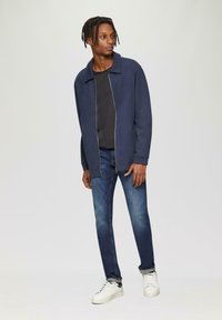 Marineblauwe zip-up jas over een zwart shirt, gecombineerd met blauwe jeans en witte sneakers. De stof is glad met een modern ontwerp en een relaxte pasvorm.