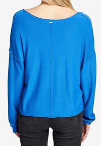 Niebieski, oversize sweter z szerokim dekoltem, opadającymi ramionami i widocznymi detalami szycia. Tył ma centralny szew i metkę blisko szyi.
