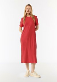 Rotes Midi-Kleid mit kurzen Ärmeln, glatter Textur und entspanntem Schnitt. Die Modells steht in einem Innenraum und trägt beigen Sneakers.