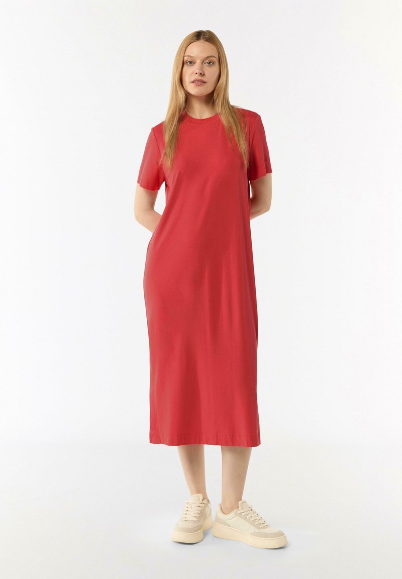 Rotes Midi-Kleid mit kurzen Ärmeln, glatter Textur und entspanntem Schnitt. Die Modells steht in einem Innenraum und trägt beigen Sneakers.