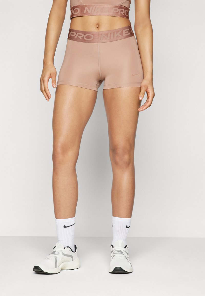 Donna che indossa pantaloni Nike Pro color nude, calze Nike bianche e scarpe da ginnastica bianche, in piedi contro uno sfondo neutro.